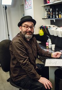 Tatér v tmavě hnědém tričku a černých kalhotách, sedící před stolem s uměleckými potřebami a skicou na papíře, usmívající se.