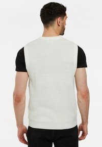 Ärmelloser cremefarbener gerippter Pullover mit rundem Halsausschnitt, figurbetontem Design und einem schwarzen T-Shirt darunter, von hinten gezeigt.