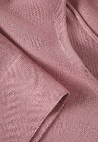 Gebreide stof in zacht, muted roze met een gestructureerde afwerking. Heeft een gladde rand en subtiele stikdetails.