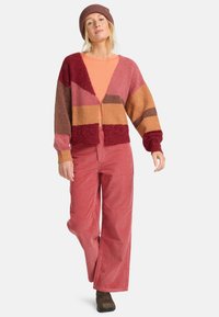 Brede gebreide cardigan in rode, bordeaux en bruine tinten met geometrische patronen; gecombineerd met een oranje top en roze corduroy broek; bruine laarzen.