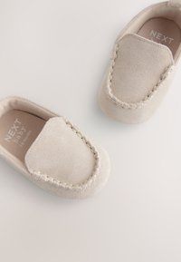Mocassins pour bébé en daim beige avec un détail cousu le long du bord et un design à bout arrondi, dotés d'une doublure intérieure douce et d'une semelle plate.