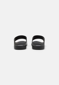Sandali in gomma nera con cinturini posteriori, suole strutturate e marchio bianco sui lati. Design semplice e piatto dallo stile minimalista.