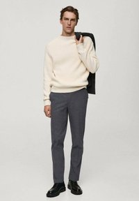 Pulover crem cu elastic, pantaloni gri și pantofi negri cu șireturi. Modelul ține un sacou negru pe umăr, stând în fața unui fundal neutru.