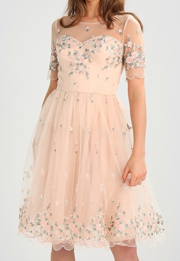 Femme portant une robe pêche transparente jusqu'aux genoux avec broderie florale et manches courtes, tenant délicatement la jupe d'une main.