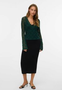 Haut en dentelle à manches longues de couleur vert foncé avec un col en forme de vagues, associé à une jupe midi noire ajustée et des sandales à talons noires.