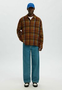 Bruin-oranje geruit overhemd, donkerblauwe spijkerbroek, zwart-witte sneakers, blauwe pet. Ontspannen pasvorm met knoopsluiting en wijde pijpen.