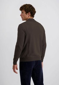 Man die naar links kijkt en een donkerbruin longsleeve polo trui draagt, samen met een marineblauwe corduroy broek, tegen een eenvoudige lichte achtergrond.