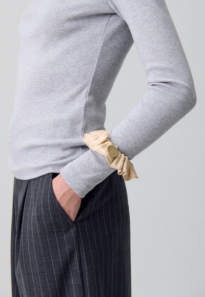 Elastico in tessuto beige con un accento dorato, indossato su una camicia grigia a maniche lunghe, abbinato a pantaloni a righe sottili scuri.