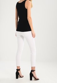 Top sans manches noir, jean skinny blanc, et sandales à talons hauts noires avec brides de cheville, vues de dos sur un fond uni.