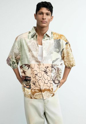 Young man with short dark hair wearing a short-sleeve patchwork floral shirt over a white undershirt and light-colored pants.