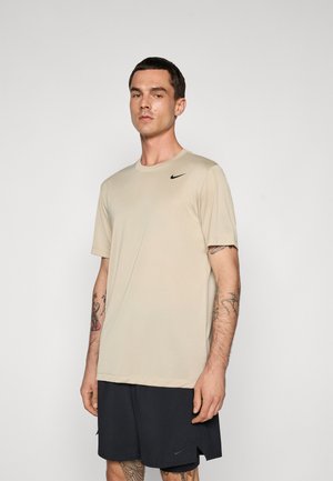 Young man with tattoos wearing beige Nike t-shirt and black Nike shorts against plain gray background.