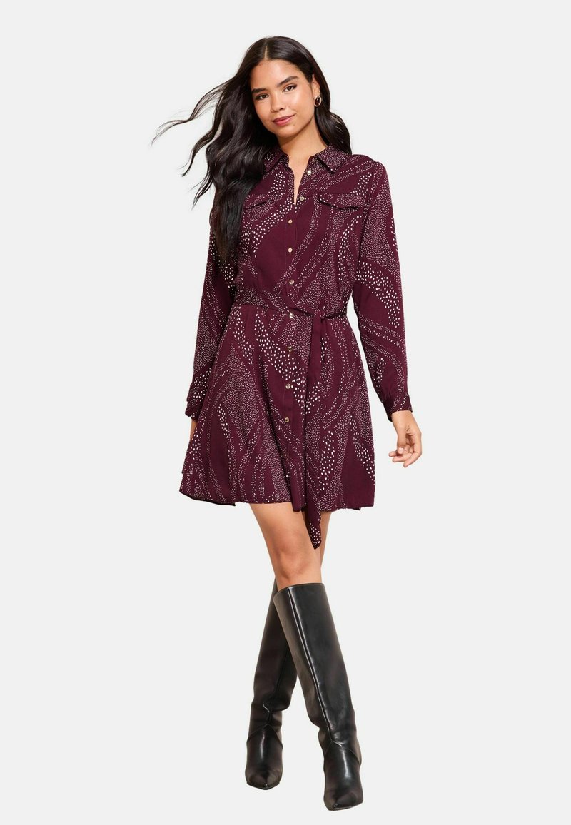 Bordeauxfarbenes Hemdkleid mit Kragen, Knopfleiste und Taillengürtel; verfügt über ein wellenförmiges weißes Muster und lange Ärmel. Kombiniert mit schwarzen knielangen Stiefeln.