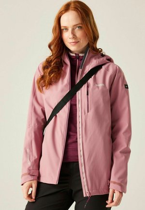 Joven con cabello rojo que lleva una chaqueta rosa para exteriores, una sudadera con cremallera morada debajo y una correa de bolso cruzado negra, sobre un fondo liso.