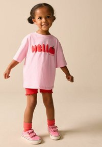 Růžové volné tričko s nápisem "Hello" a grafikou rajčete, doplněné červenými šortkami a růžovými teniskami se suchými zipy a barevnými detaily.