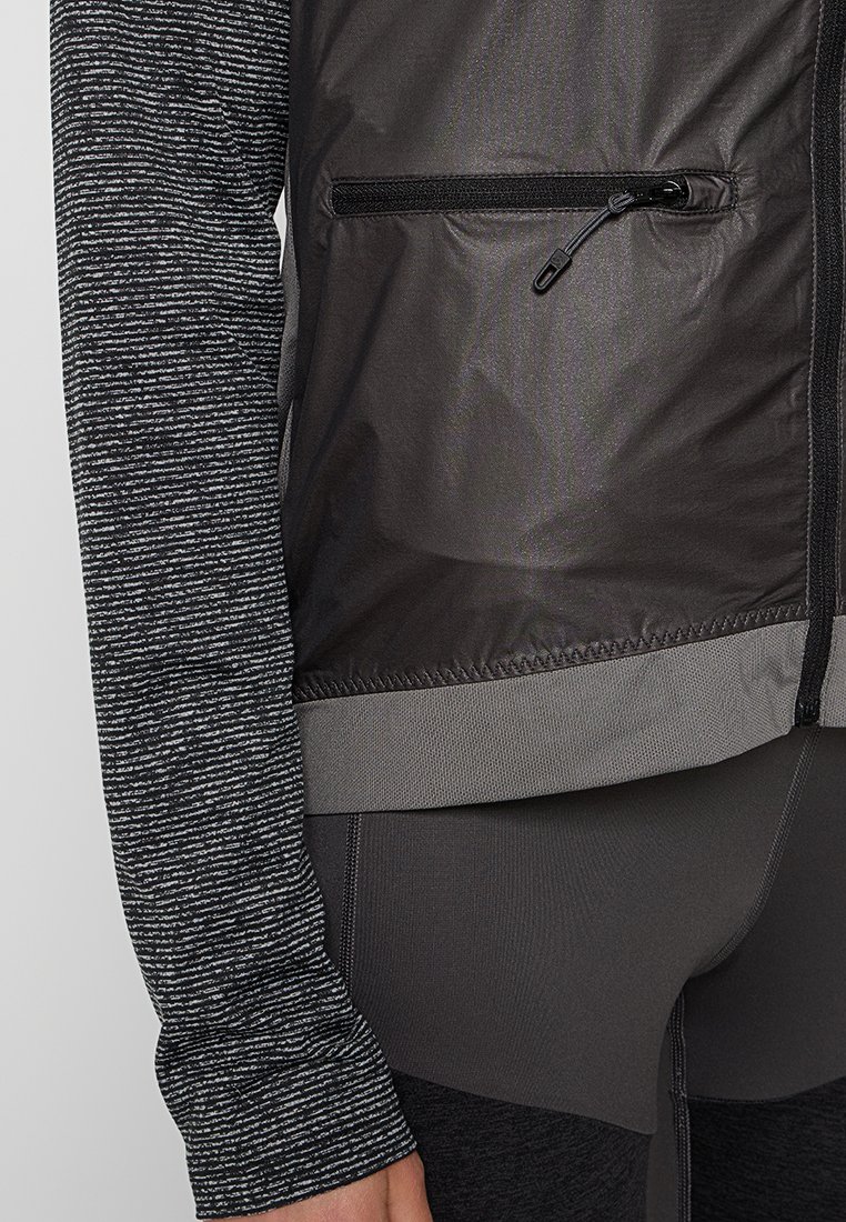 Nahaufnahme einer Person, die eine schwarze und graue Reißverschlussweste, einen grauen Bund und ein schwarzes und graues Langarmshirt sowie Leggings trägt.