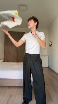 Fiatal férfi fehér ingben és fekete nadrágban egy fehér inget dob fel, miközben egy fekete tárgyat tart a kezében egy hálószobában.