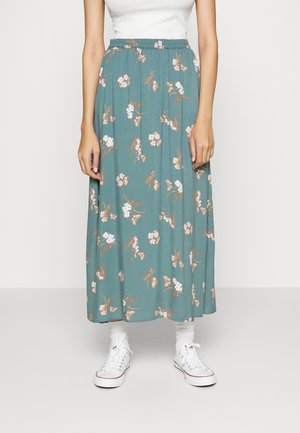 Femme portant une jupe midi fleurie bleu sarcelle avec une taille élastique, des baskets blanches, des chaussettes blanches et un haut blanc sans manches.