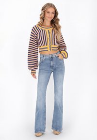 Gehaakt vest met horizontale strepen in paars en geel, voorzien van grote knopen. Gecombineerd met hooggetailleerde, uitlopende lichtblauwe jeans.
