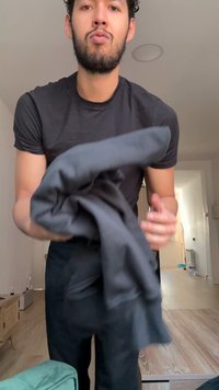 Homme aux cheveux bouclés foncés et à la barbe, portant une chemise noire, tenant et pliant un vêtement gris foncé dans une pièce lumineuse.