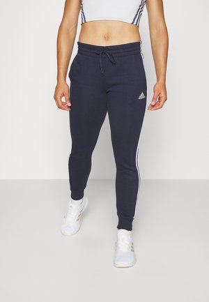 Persoon die marineblauwe Adidas joggers met witte zijstrepen en witte sportschoenen draagt, staand tegen een effen achtergrond.