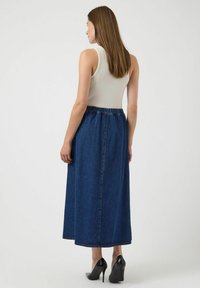 Femme de dos portant un haut sans manches blanc, une longue jupe en denim bleue et des chaussures à talons noirs sur un fond uni.