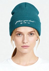 Cappellino a coste in maglia verde acqua con testo ricamato bianco "segui il tuo cuore" con design di freccia e cuore. Tessuto morbido ed elastico.