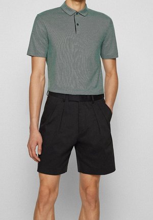 Grünes karierter Poloshirt mit Kragen, kombiniert mit schwarzen gefalteten Shorts und einem schwarzen Gürtel. Glatte Stofftextur im gesamten Outfit.