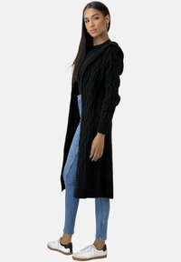 Schwarzer Strickcardigan mit Kapuze, langen Ärmeln und offener Frontanlage, kombiniert mit hellblauen Skinny-Jeans und weißen Sneakern.