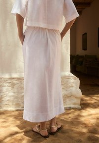 Robe en lin blanc fluide avec une taille cintrée, des manches courtes et des poches sur les côtés. Associée à des sandales plates avec des accents dorés. Texture douce.