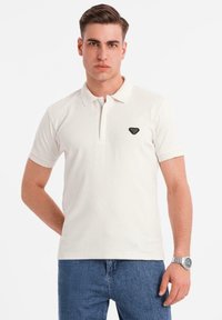 Vit polo skjorta med korta ärmar, knäppning framtill och en svart triangel-logga på bröstet. Bärs tillsammans med blå denimjeans.