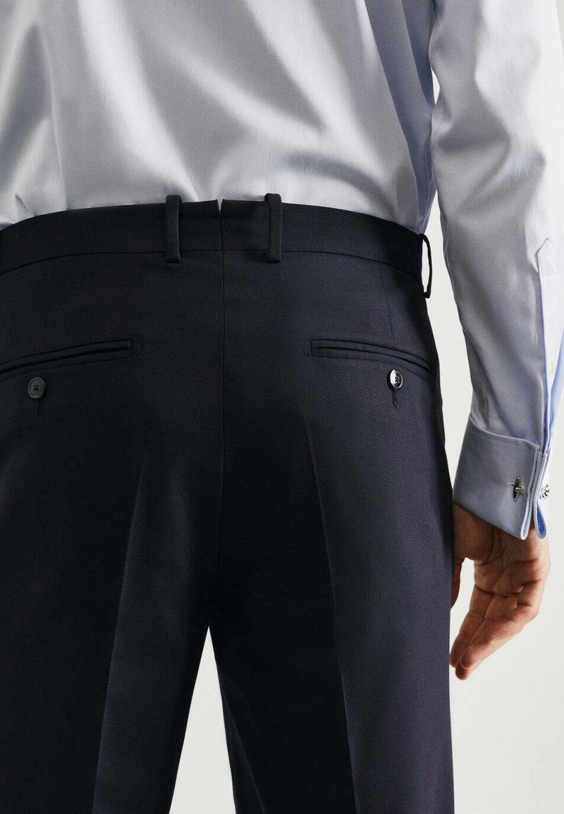 Pantalons bleu marine foncé avec une finition texturée, dotés de deux poches et de boutons. Portés avec une chemise bleu clair, montrant un tissu lisse.