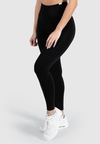 Černé legíny s vysokým pasem a hladkým, elastickým materiálem. Nosené s bílými teniskami, které mají vzorovaný akcent na ponožkách.