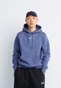 Felpa in pile blu navy con tasca frontale tipo kangaroo e lacci. Presenta un piccolo logo Nike verde neon sul petto. Pantaloni sudati neri con logo.