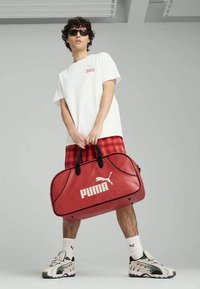 Red Puma duffel bag with black handles, featuring white logo and accents. Model wears white tee, red plaid shorts, and sneakers.