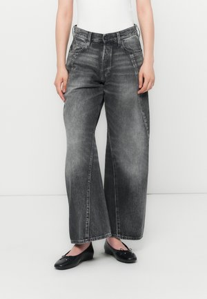 Femme portant un jean noir délavé taille haute à jambes larges et des ballerines noires, debout devant un fond blanc uni.