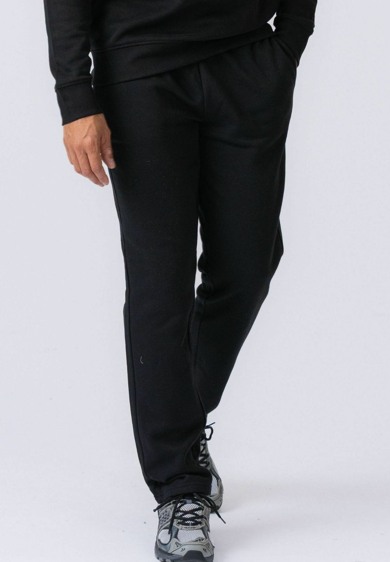 Person som har på seg svarte treningsbukser og grå joggesko, står med den ene hånden i lomma og det ene benet lett bøyd.