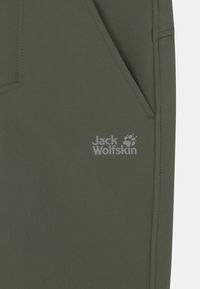 Housut oliivinvihreää väriä, jotka on valmistettu sileästä kankaasta. Niissä on sivutasku ja harmaalla painatuksella varustettu "Jack Wolfskin" -logo, jossa on tassunjälki yksityiskohtana.