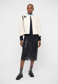 Balta sutrumpinta apsiausta su juodais gėlių siuvinėjimais virš tamsaus megztinio, derinama su prigludusia juoda blizgiu sijonu ir juodais loaferiais su kojinėmis.