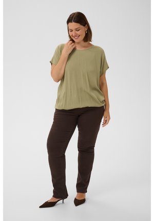 Vrouw met kort bruin haar, die een losse lichtgroene top, donkerbruine broek en donkerbruine hakken draagt, staand en glimlachend met hand bij het gezicht.