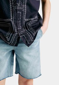 Camisa negra de botones con patrones en blanco de paisley y geométricos; combinada con pantalones cortos de mezclilla azul claro, con dobladillo deshilachado y un corte relajado.