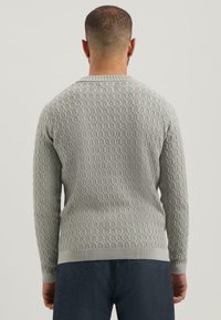 Man in een lichtgrijze, gestructureerde gebreide trui en donkere broek, van achteren afgebeeld tegen een effen lichte achtergrond.