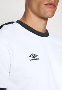 Balts T-krekls ar melnu apakšmalu un plecu akcentiem. Uz krūtīm ir melns "UMBRO" logo. Gluda auduma tekstūra.