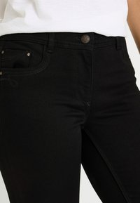 Vaqueros de mezclilla negra con un diseño ajustado, que cuentan con cierre de botones, bolsillos delanteros y traseros, y detalles de costura sutiles.