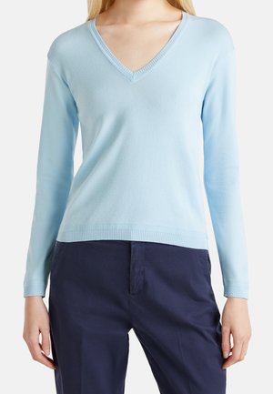 Kobieta ubrana w jasnoniebieski sweter z długim rękawem i dekoltem w serek oraz ciemnogranatowe spodnie, pokazana od szyi do bioder na białym tle.