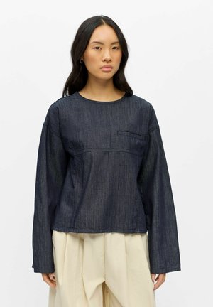 Frau mit langen dunklen Haaren, die ein dunkles Denim-Oberteil mit langen Ärmeln und lockere beige plissierte Hosen vor einem schlichten weißen Hintergrund trägt.