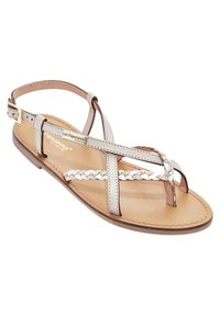 Sandalo piatto con suola in pelle beige, dotato di cinturini intrecciati in marrone chiaro e argento. Una cinghia regolabile alla caviglia offre stabilità.