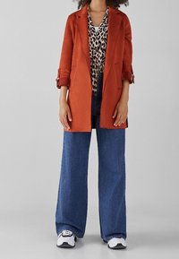 Abrigo entallado de color óxido sobre una blusa con estampado de leopardo, combinado con jeans de pierna ancha en azul y zapatillas en blanco y negro.