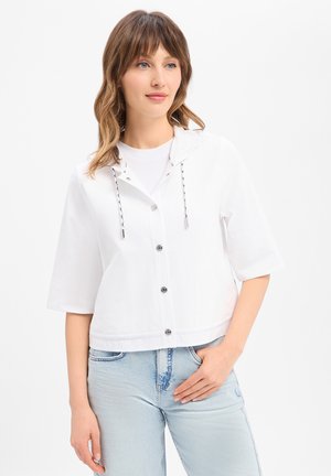 Jeune femme aux cheveux châtain clair, portant un sweat à capuche blanc boutonné à manches courtes et un jean bleu clair, debout avec une main dans la poche.