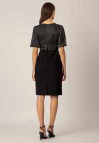 Femme debout, portant une robe noire avec un haut texturé et scintillant, et des manches courtes bouffantes, accompagnée de talons hauts noirs à lanières.