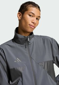 Šedá športová bunda s vysokým golierom, zipsom a kontrastnými matnými a lesklými panelmi. Obsahuje reflexný detail s logom.
